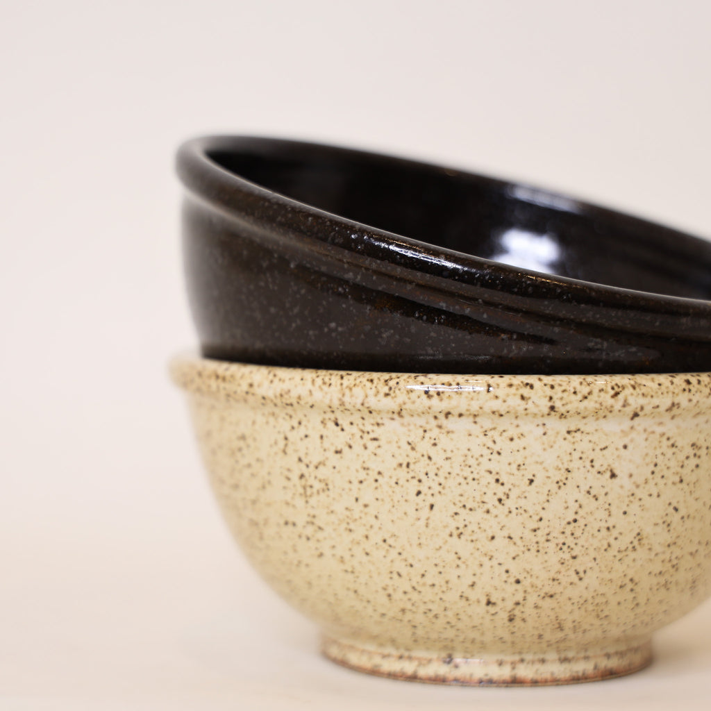 Two ceramic bowls, one black and one beige speckled, stacked on a light background.