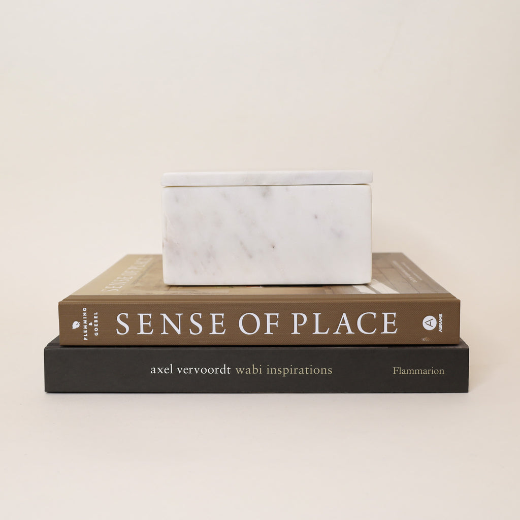 White marble box stacked on top of two books titled 'Sense of Place' and 'axel vervoordt wabi inspirations' on a beige background.