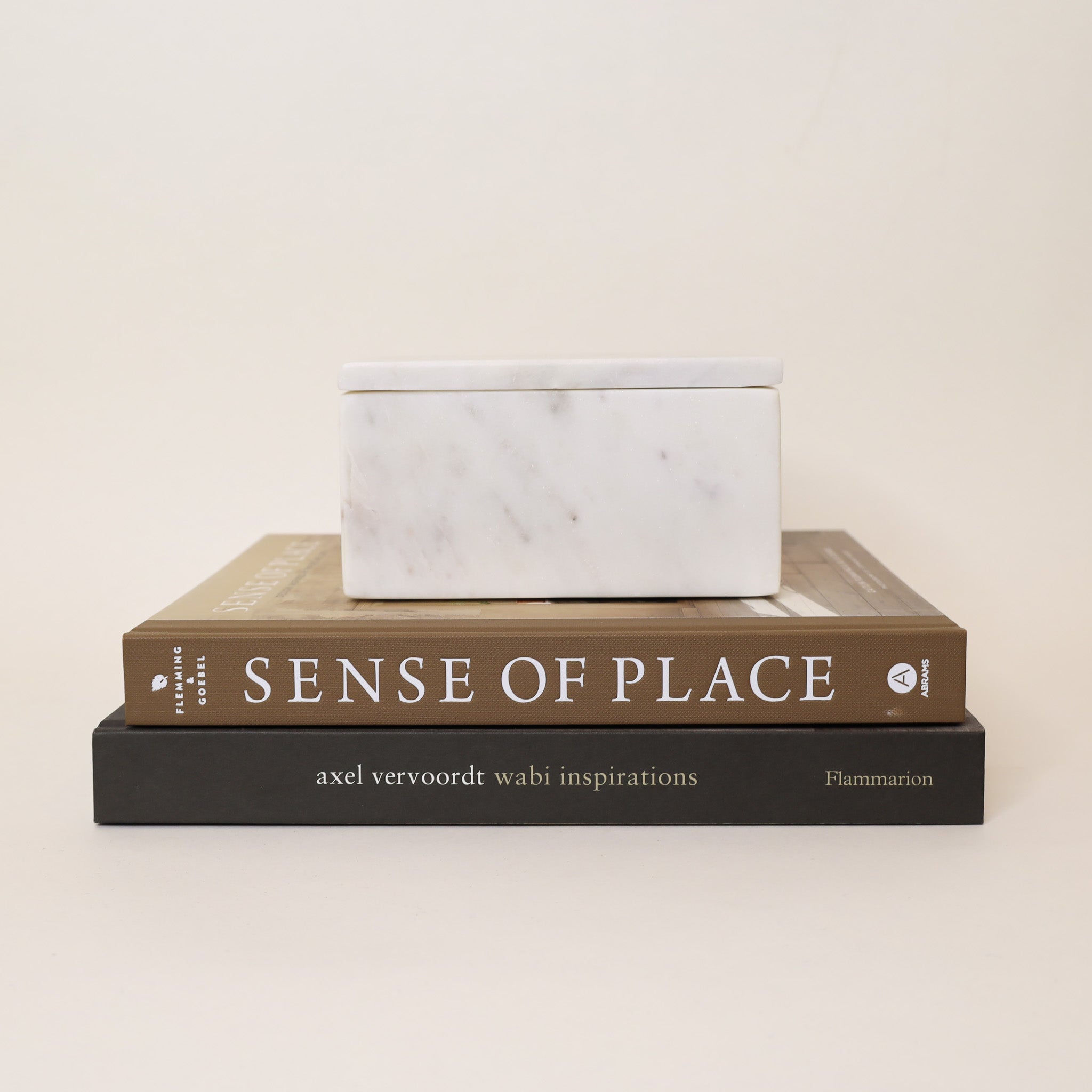 White marble box stacked on top of two books titled 'Sense of Place' and 'axel vervoordt wabi inspirations' on a beige background.