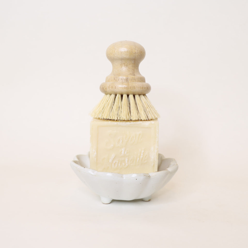 Dish brush with wooden handle and bristles on a white soap bar, placed on a white ceramic bowl against a white background.
