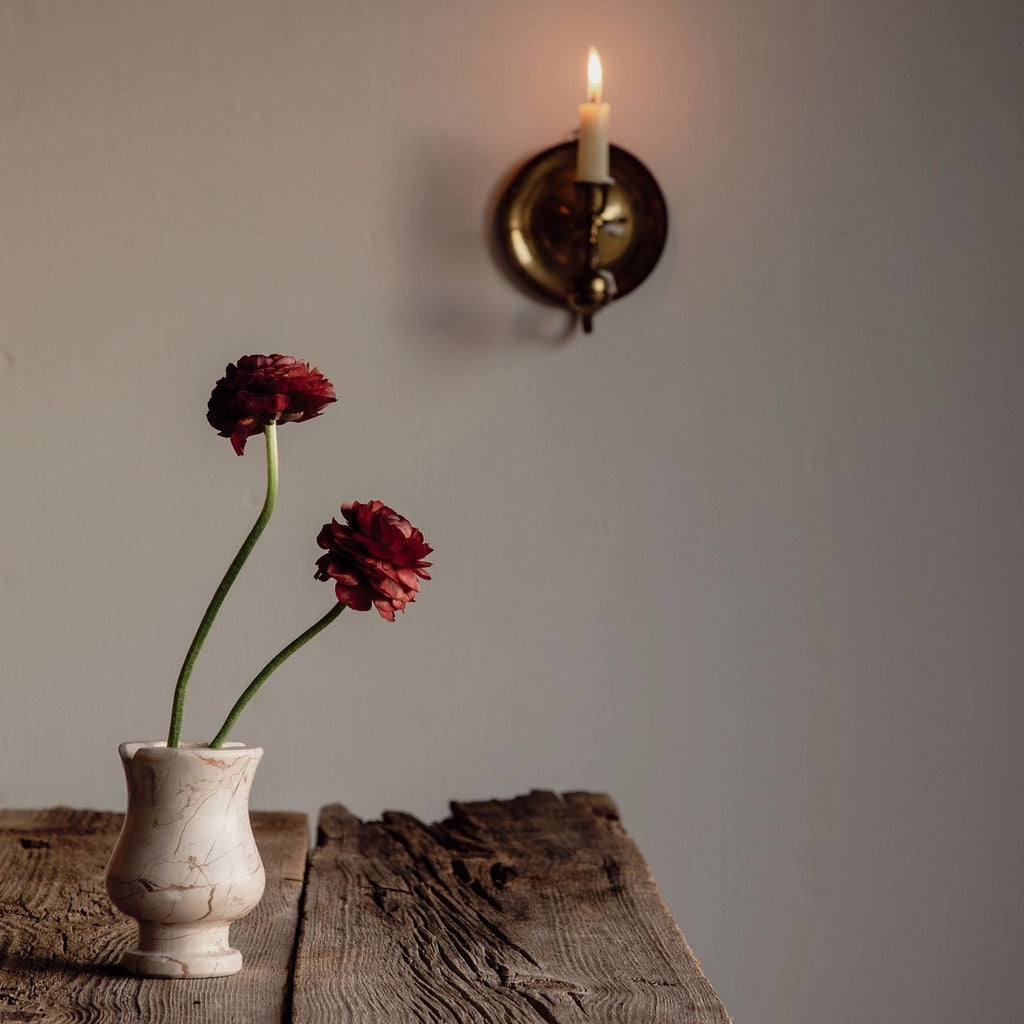 Two red flowers in a white vase on a wooden surface with a wall sconce in the background.