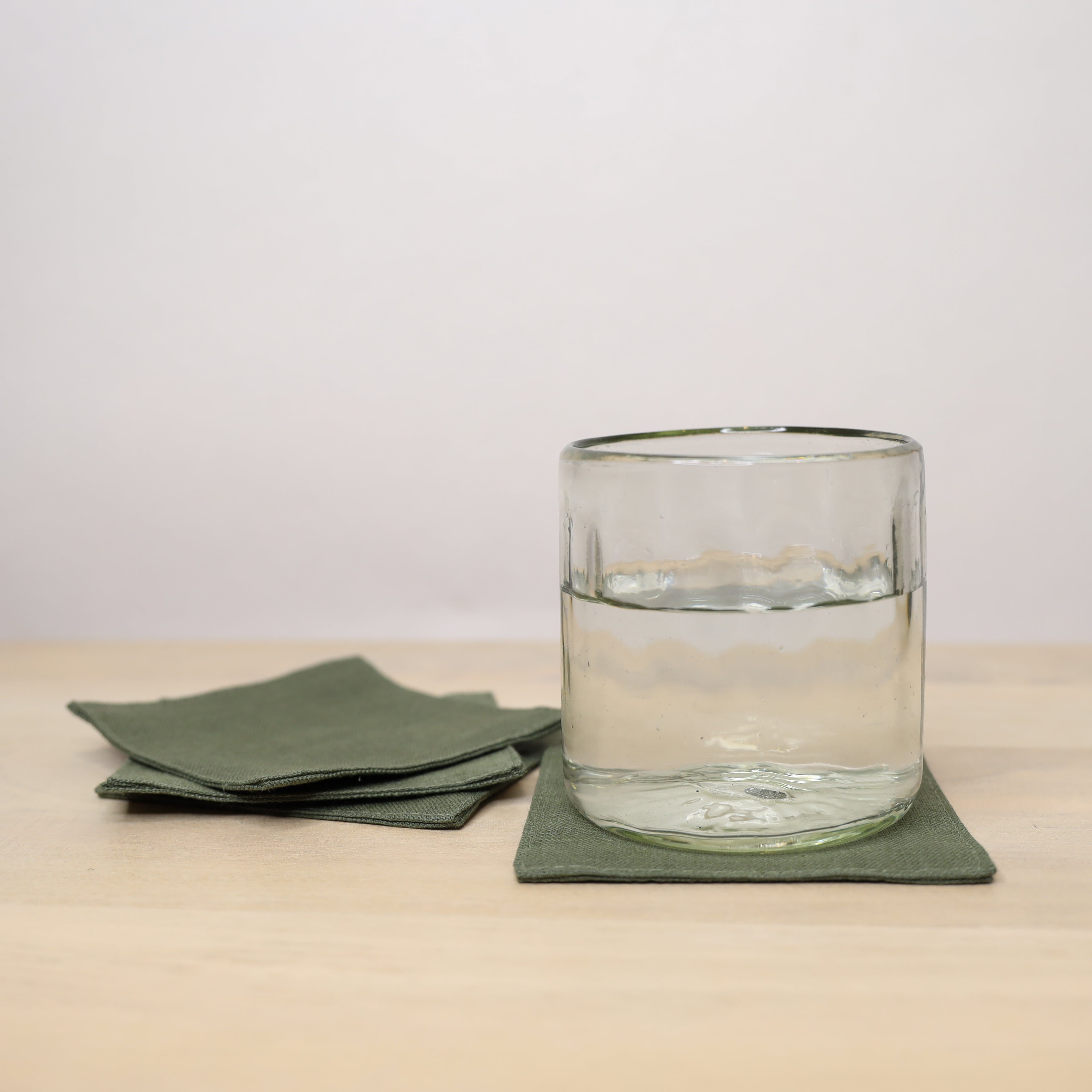 Clear glass with water on a green coaster, surrounded by green napkins on a beige surface.