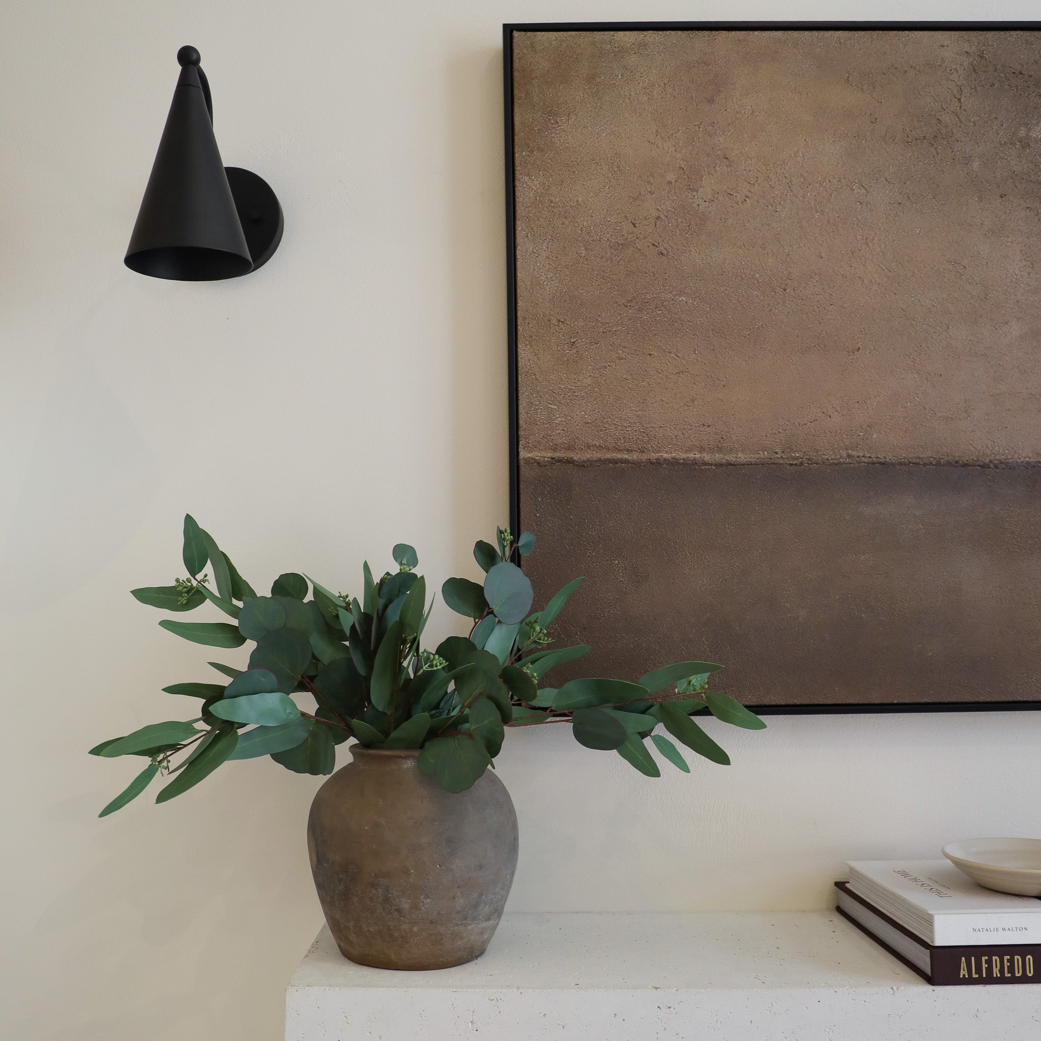 Vase with green leaves on a shelf next to a framed abstract painting.